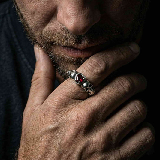 Detalle de anillo artesanal de hombre con calaveras laterales y corazón granate en plata de ley pulida.