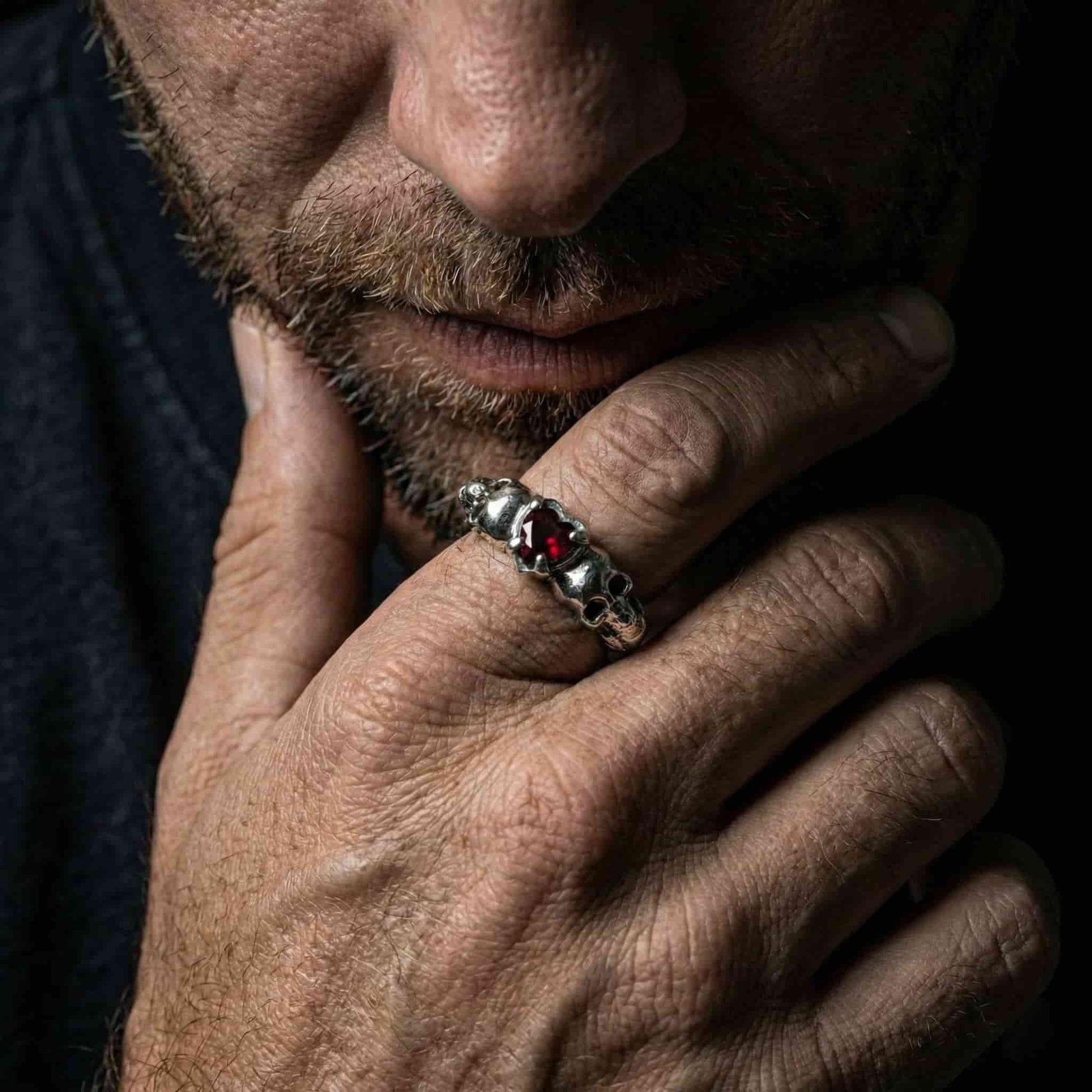 Detalle de anillo artesanal de hombre con calaveras laterales y corazón granate en plata de ley pulida.
