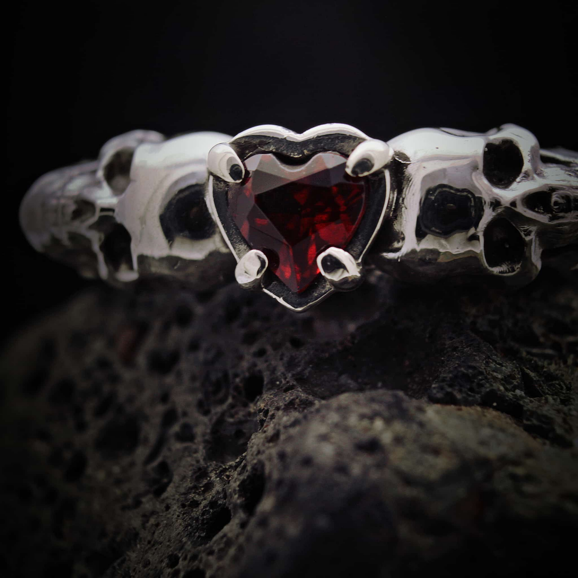 Alianza de boda gótica en plata con diseño de calaveras y corazón rojo granate.