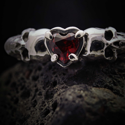 Alianza de boda gótica en plata con diseño de calaveras y corazón rojo granate.