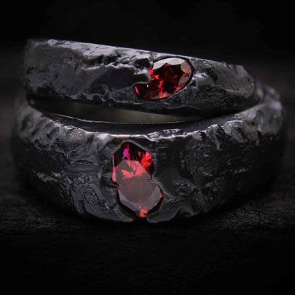 Primer plano de estudio de un set de dos alianzas de boda artesanales forjadas en plata negra oxidada con tres circonitas rojas ovaladas engastadas.