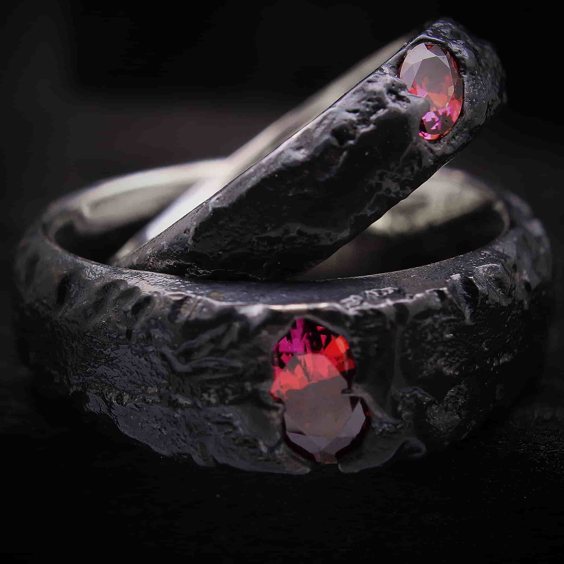 Detalle de la alianza de 7mm de ancho de plata negra forjada con dos circonitas rojas ovaladas visibles engastadas de forma orgánica.