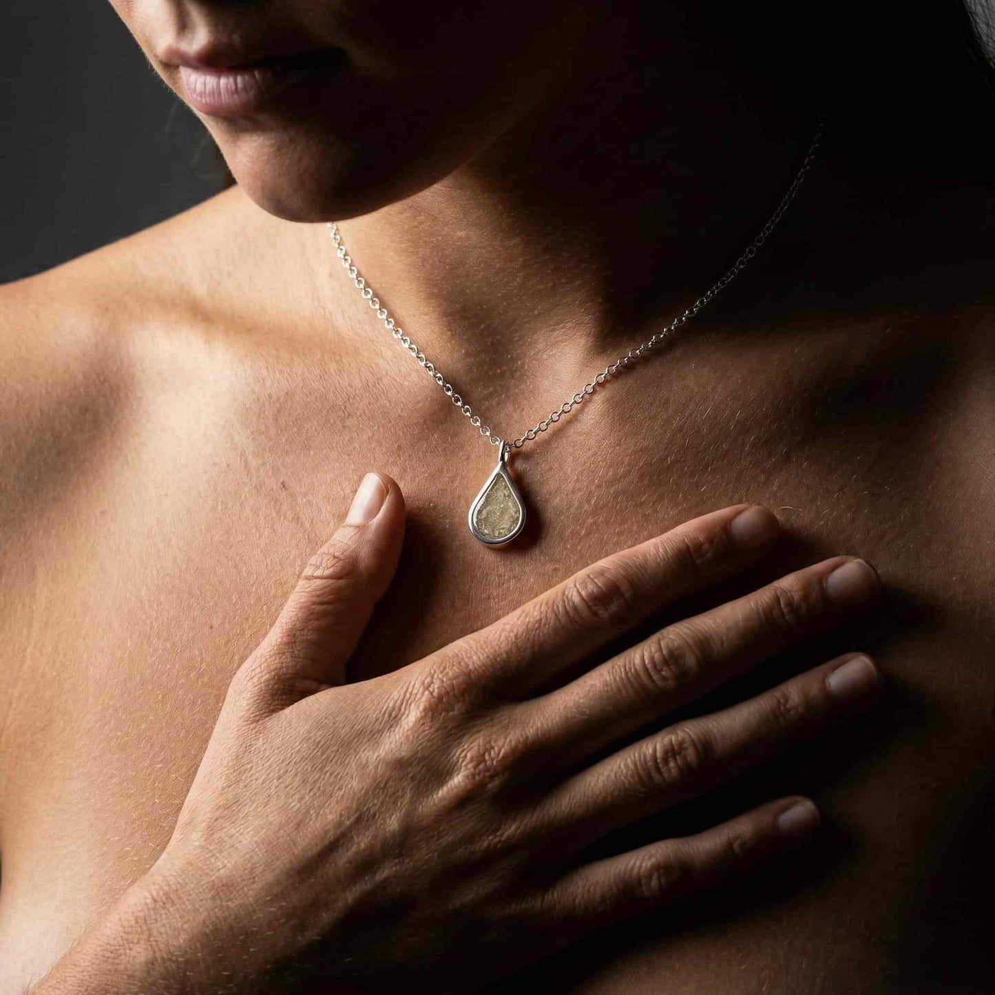 Primer plano de un colgante de plata en forma de lágrima que contiene cenizas de recuerdo, colgado del cuello de una persona con una mano apoyada suavemente sobre el pecho.